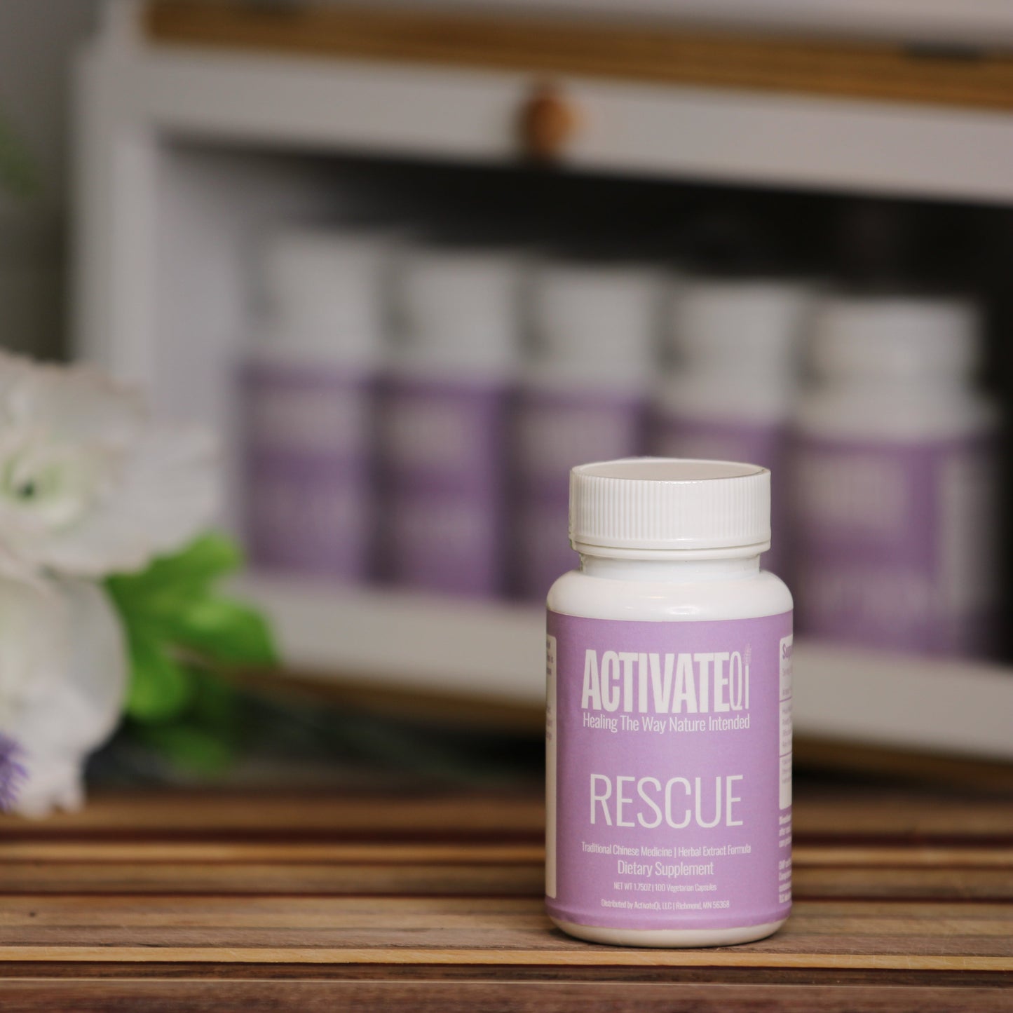 Bottle of ActivateQi Rescue on a wooden surface with flowers and more bottles in the background.