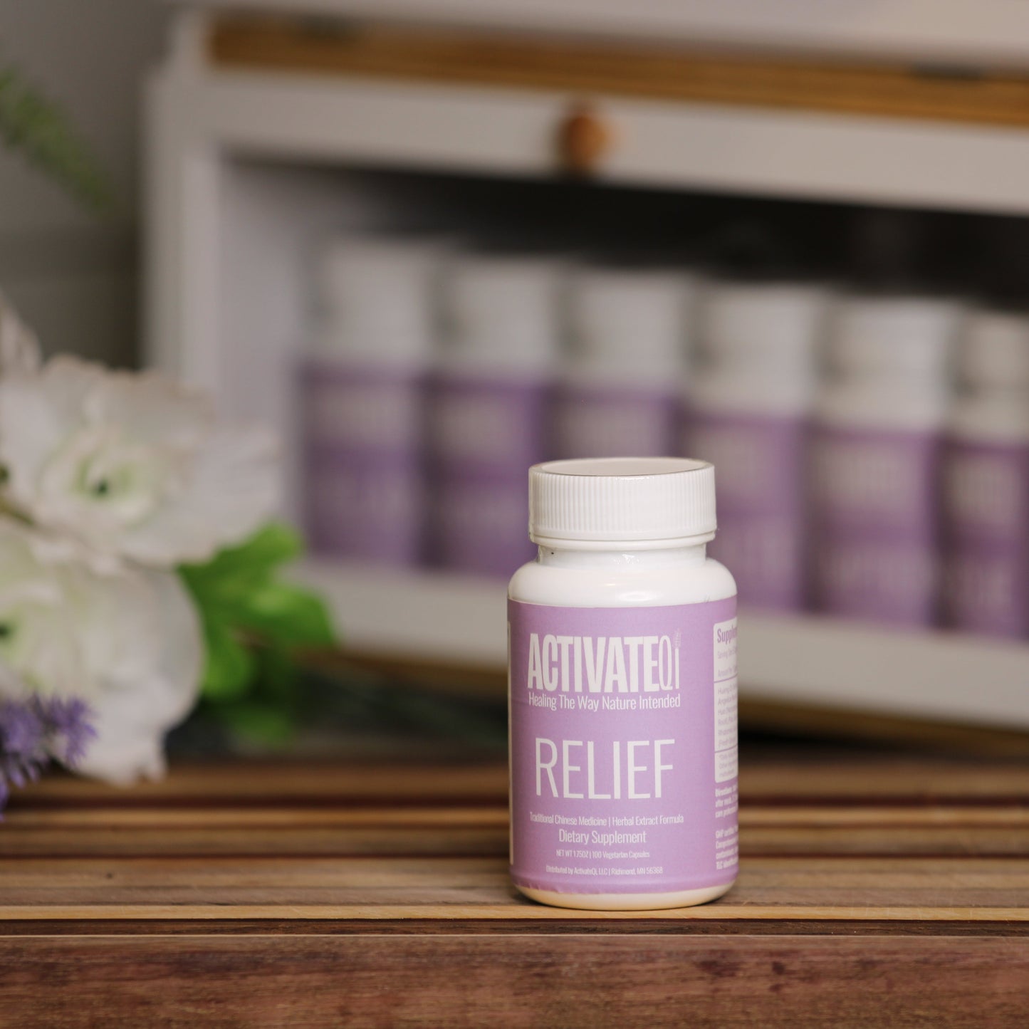 Purple bottle labeled 'ActivateQi Relief' on a wooden surface with flowers and more bottles in the background.