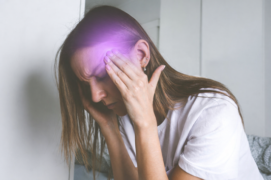 Woman holding her temples with eyes closed in discomfort, with a soft purple glow highlighting the side of her forehead to represent head pain or a migraine.