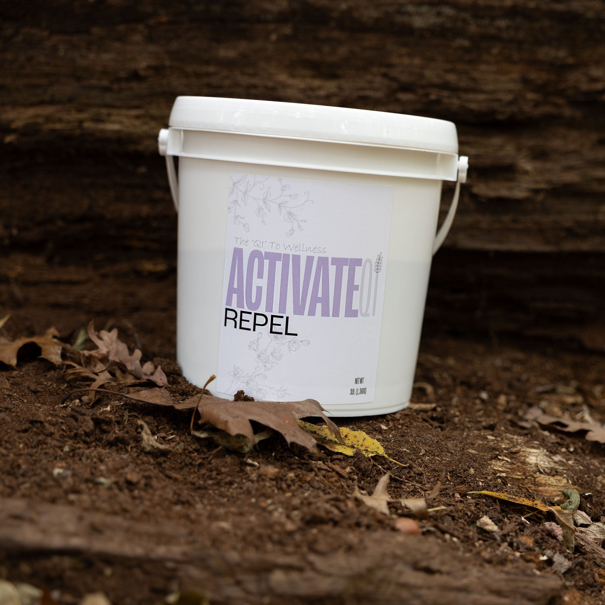 White bucket labeled ActivateQi Repel on a dirt surface with leaves.