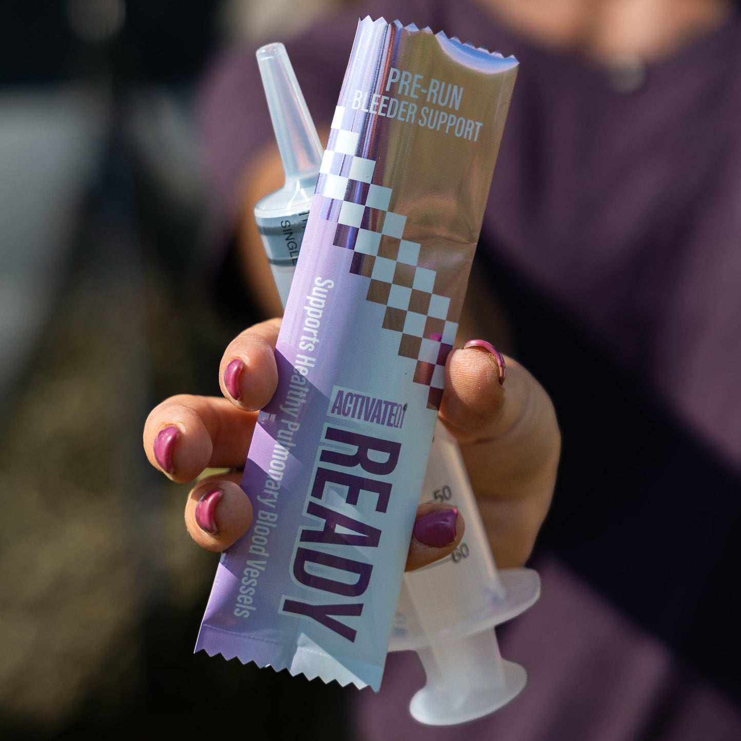 A hand with pink nail polish holds a tube of Ready Pre-Run EIPH Support alongside a syringe in front of a blurred person wearing a purple top.