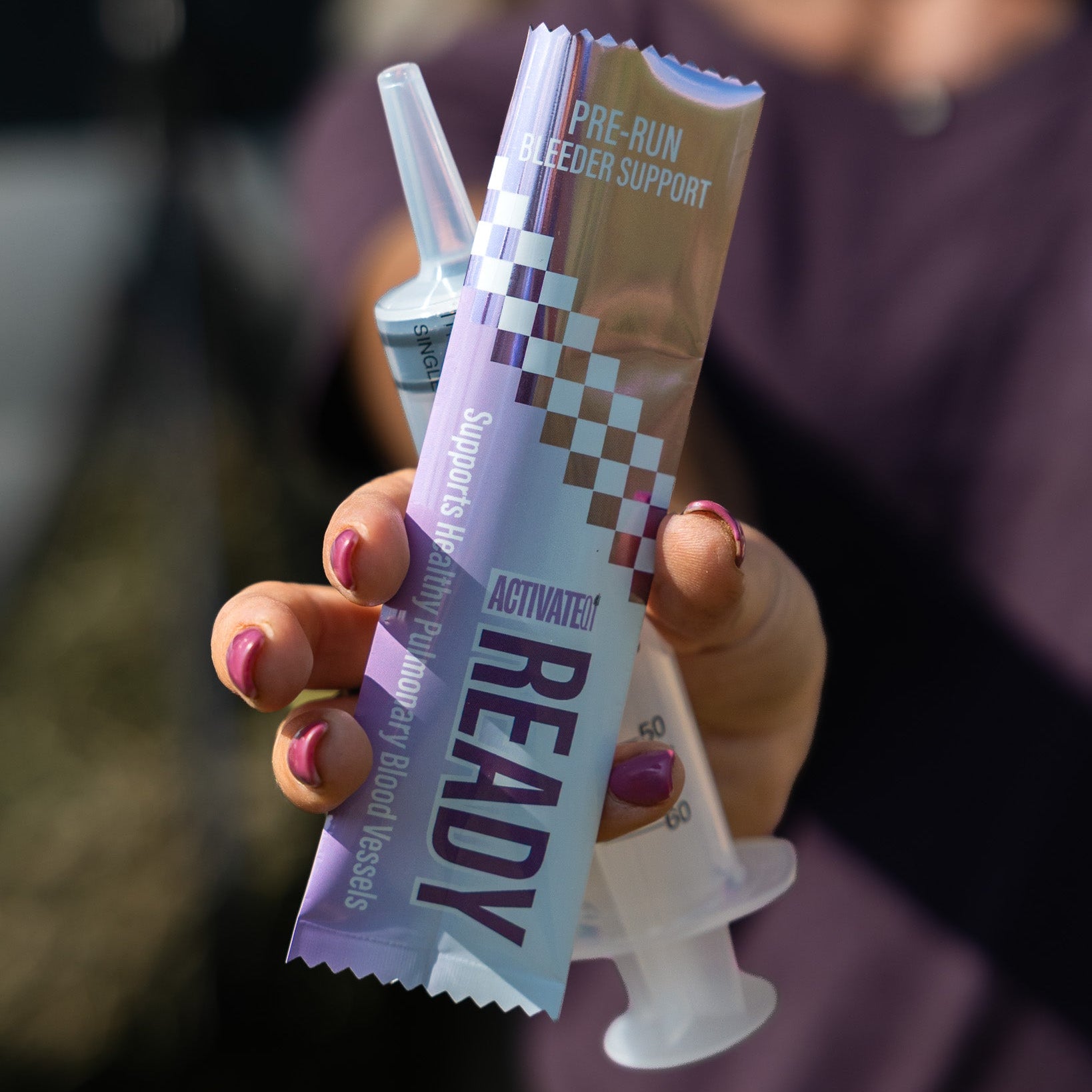 A hand with pink nail polish holds a tube of Ready Pre-Run EIPH Support alongside a syringe in front of a blurred person wearing a purple top.