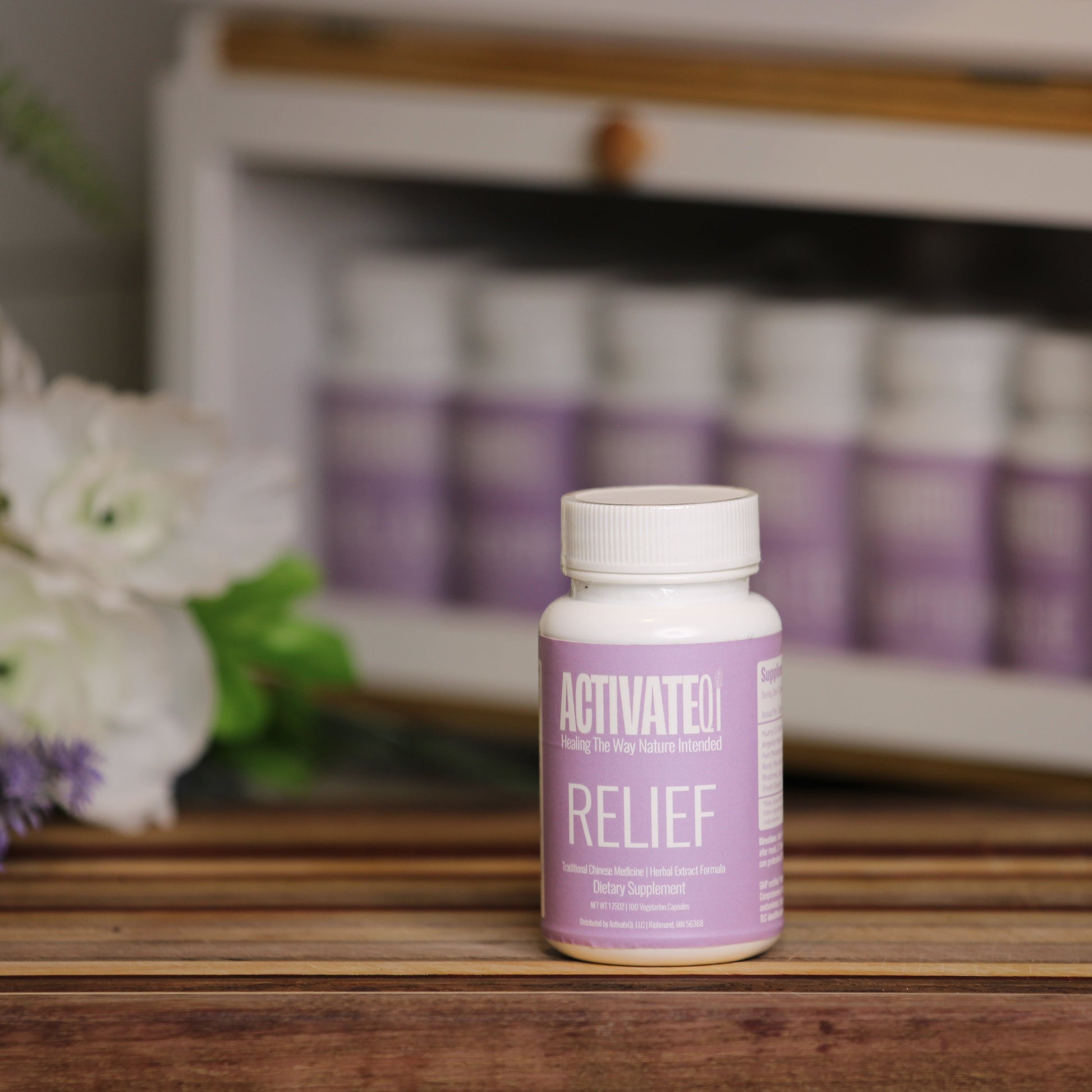 Purple bottle labeled 'ActivateQi Relief' on a wooden surface with flowers and more bottles in the background.